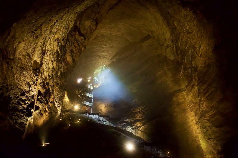 Looking up inside a vast cave chamber lit by warm lamps, with a narrow staircase and railing winding along the rock wall toward a shaft of daylight