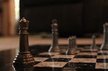 A close-up view of a chessboard with metallic chess pieces, prominently featuring a bronze queen in the foreground. The background is softly blurred, accentuating the focus on the queen. The chessboard squares are black and white, adding a sense of strategy and contemplation to the scene.