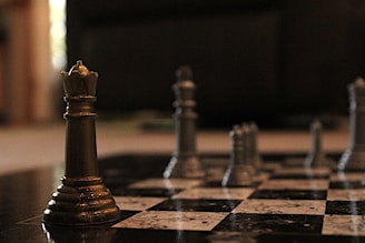 A close-up view of a chessboard with metallic chess pieces, prominently featuring a bronze queen in the foreground. The background is softly blurred, accentuating the focus on the queen. The chessboard squares are black and white, adding a sense of strategy and contemplation to the scene.