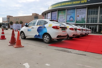 A row of white cars with blue branding parked on a red carpet near a building showcasing advertisements related to a renewable energy expo. The scene includes several orange traffic cones placed along the side.