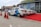 A row of white cars with blue branding parked on a red carpet near a building showcasing advertisements related to a renewable energy expo. The scene includes several orange traffic cones placed along the side.