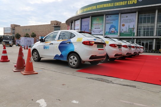 A row of white cars with blue branding parked on a red carpet near a building showcasing advertisements related to a renewable energy expo. The scene includes several orange traffic cones placed along the side.