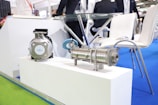 A close-up view of industrial equipment placed on a white platform in a professional exhibition setting. The floor is covered with bright blue and green carpeting, and there are modern white chairs around a glass table in the background with some people in business attire.