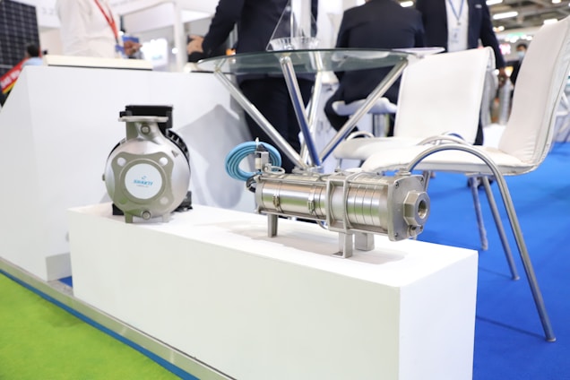 A close-up view of industrial equipment placed on a white platform in a professional exhibition setting. The floor is covered with bright blue and green carpeting, and there are modern white chairs around a glass table in the background with some people in business attire.