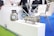A close-up view of industrial equipment placed on a white platform in a professional exhibition setting. The floor is covered with bright blue and green carpeting, and there are modern white chairs around a glass table in the background with some people in business attire.