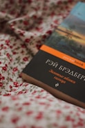 A book with a dark cover featuring Cyrillic text, lying on a floral fabric. The fabric has a white background with red rose patterns. The book has a strip on top with an orange label.
