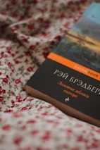A book with a dark cover featuring Cyrillic text, lying on a floral fabric. The fabric has a white background with red rose patterns. The book has a strip on top with an orange label.