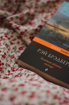 A book with a dark cover featuring Cyrillic text, lying on a floral fabric. The fabric has a white background with red rose patterns. The book has a strip on top with an orange label.