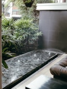 A modern outdoor space features a small rectangular water fountain with jets creating small splashes. Surrounding the fountain are lush green plants and a dark tiled wall, providing a serene and refreshing ambiance. To the right, a brown leather sofa suggests a comfortable seating area.