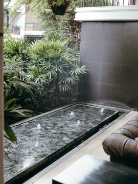 A modern outdoor space features a small rectangular water fountain with jets creating small splashes. Surrounding the fountain are lush green plants and a dark tiled wall, providing a serene and refreshing ambiance. To the right, a brown leather sofa suggests a comfortable seating area.