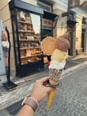 A hand holding a large ice cream cone with three different scoops, each a different color, topped with a waffle disc. The ice cream is held near a gelateria storefront with a display of desserts and pastries inside. The street is paved with cobblestones and there is a sign offering a glimpse of the shop's name and branding.