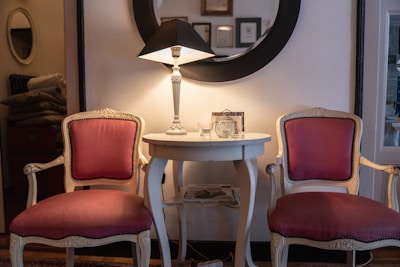 A cozy interior scene featuring two elegant chairs with ornate woodwork and pink upholstery, positioned around a round, white table. A table lamp with a black shade casts a warm glow, and a large round mirror hangs on the wall behind, reflecting part of the room. Various decorative items, including framed pictures and a small clock, adorn the table and wall.