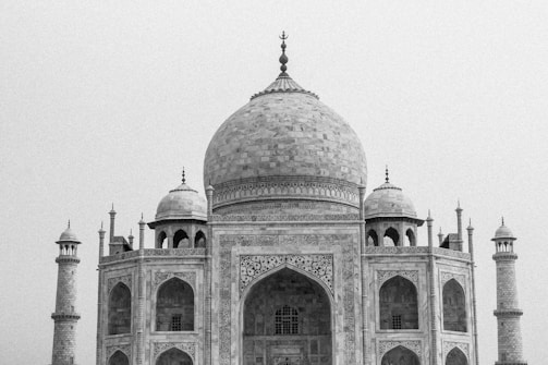 A detailed architectural view of a large, intricate marble building featuring a central dome flanked by four smaller domes and tall minarets. The structure displays ornate carvings and arches, showcasing a blend of symmetrical and elegant design elements typical of Mughal architecture.
