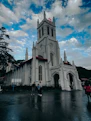 A large, white Gothic-style church stands with a prominent red cross at the top. The building features tall arched windows and elaborate details along the facade. Three people are gathered in conversation in front of the church on a wet pavement. The sky above is partly cloudy, with patches of blue visible.