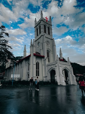 A large, white Gothic-style church stands with a prominent red cross at the top. The building features tall arched windows and elaborate details along the facade. Three people are gathered in conversation in front of the church on a wet pavement. The sky above is partly cloudy, with patches of blue visible.