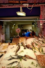 A fish market stall displays a variety of fish on a bed of ice. Several types of fish, including whole fish and fillets, are neatly arranged with sprigs of fresh herbs. In the background, a scale is visible, and three people are engaged in conversation.
