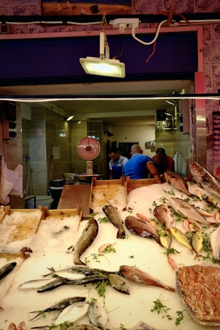 A fish market stall displays a variety of fish on a bed of ice. Several types of fish, including whole fish and fillets, are neatly arranged with sprigs of fresh herbs. In the background, a scale is visible, and three people are engaged in conversation.