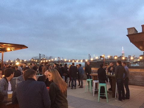 A rooftop gathering during dusk with groups of people socializing. String lights create a warm ambiance against the backdrop of a city skyline. The sky is overcast, and individuals are seated and standing, some holding drinks.