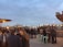 A rooftop gathering during dusk with groups of people socializing. String lights create a warm ambiance against the backdrop of a city skyline. The sky is overcast, and individuals are seated and standing, some holding drinks.