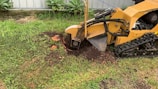 A yellow stump grinding machine is actively grinding a tree stump in a grassy area. The machine has a large rotating blade, visible in motion, cutting through the stump. There are patches of soil and wood chips scattered around the grinding area. In the background, there is a corrugated metal fence and green foliage.
