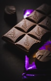 A close-up of a chocolate bar with several pieces visible, resting on a partially visible purple wrapper. The surface of the chocolate appears smooth and slightly glossy, with a few decorative swirls. Two additional small chocolate pieces are placed nearby.