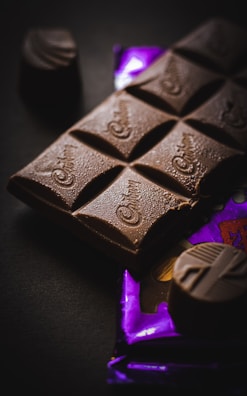 A close-up of a chocolate bar with several pieces visible, resting on a partially visible purple wrapper. The surface of the chocolate appears smooth and slightly glossy, with a few decorative swirls. Two additional small chocolate pieces are placed nearby.