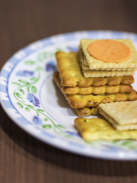 A stack of square crackers is arranged neatly on a decorative plate. The crackers appear layered, with one cracker having a slice of cheese on top. The plate features a blue and white design with floral patterns, and the background surface is a wooden table.