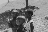 Three young children are standing outdoors, appearing to be engaged in conversation or play. The setting is sunny, with visible shadows and a tree in the background. The children display expressions of joy and curiosity.