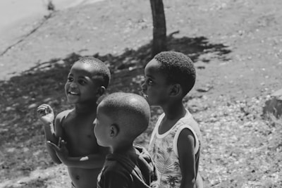 Three young children are standing outdoors, appearing to be engaged in conversation or play. The setting is sunny, with visible shadows and a tree in the background. The children display expressions of joy and curiosity.