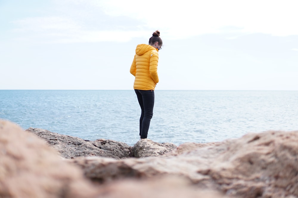 a woman in a yellow jacket standing on a rock by the ocean