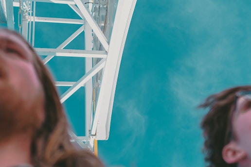 An abstract perspective with two partially visible faces, one on each side of the image in the foreground. The background consists of a structural steel construction with a bright blue sky. The image is slightly out of focus, giving it a dreamy, surreal quality.