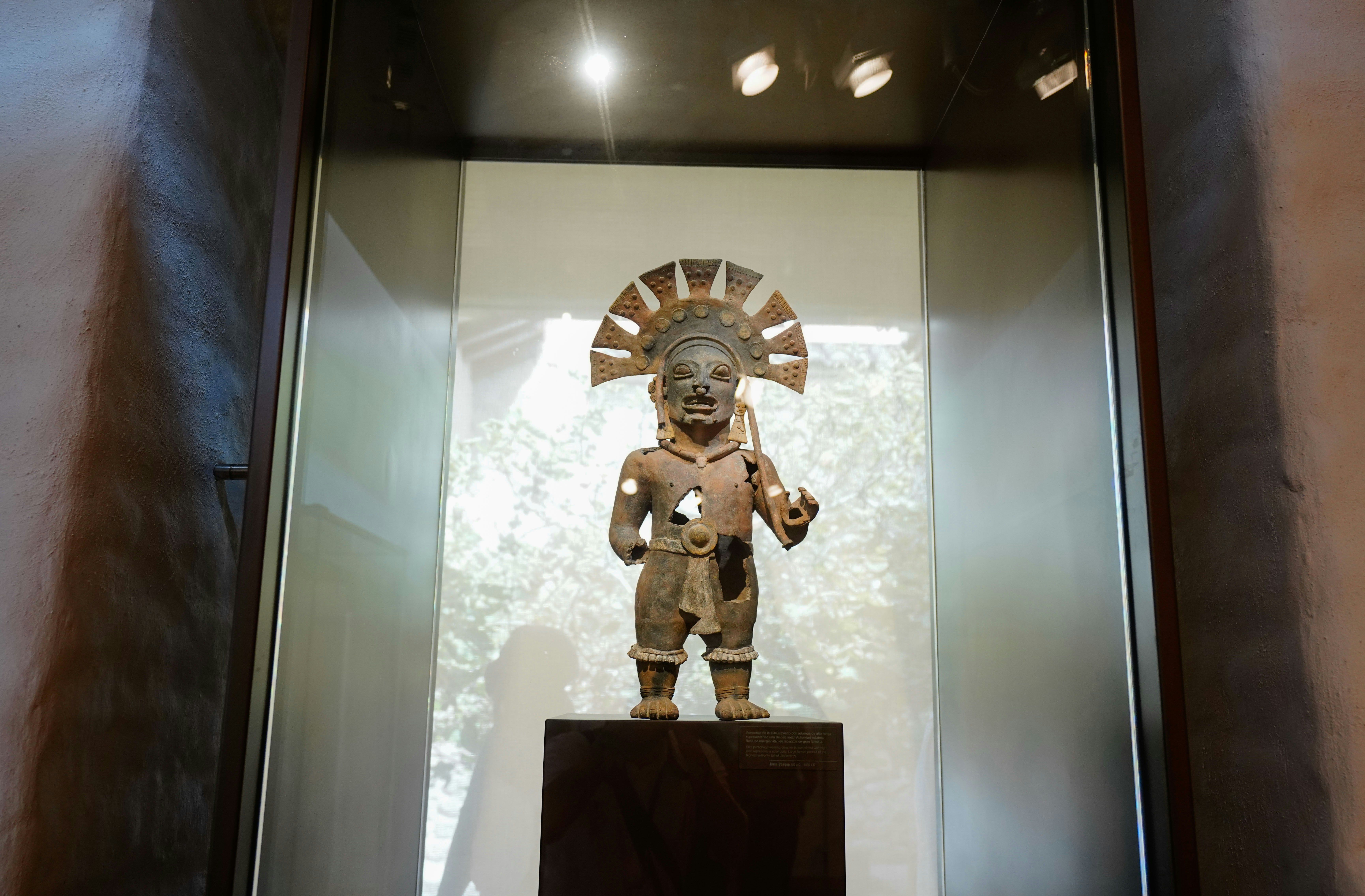 Tête en bronze dorée aux traits royaux et coiffe cérémonielle, éclairée dramatiquement dans vitrine de musée sombre