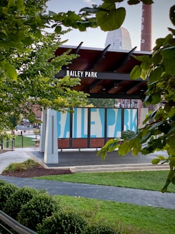 A park sign reading 'Bailey Park' is centered, surrounded by lush green trees and bushes. The park features a modern structure with a wooden and metal design. Behind the sign, a tall brick chimney is visible, and a building with blue markings is partially obscured by foliage. The scene is peaceful with a few pedestrians in the background and bright, natural lighting.