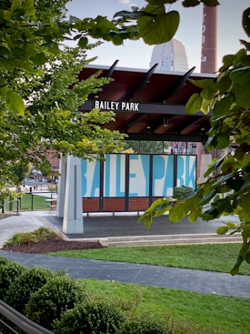 A park sign reading 'Bailey Park' is centered, surrounded by lush green trees and bushes. The park features a modern structure with a wooden and metal design. Behind the sign, a tall brick chimney is visible, and a building with blue markings is partially obscured by foliage. The scene is peaceful with a few pedestrians in the background and bright, natural lighting.