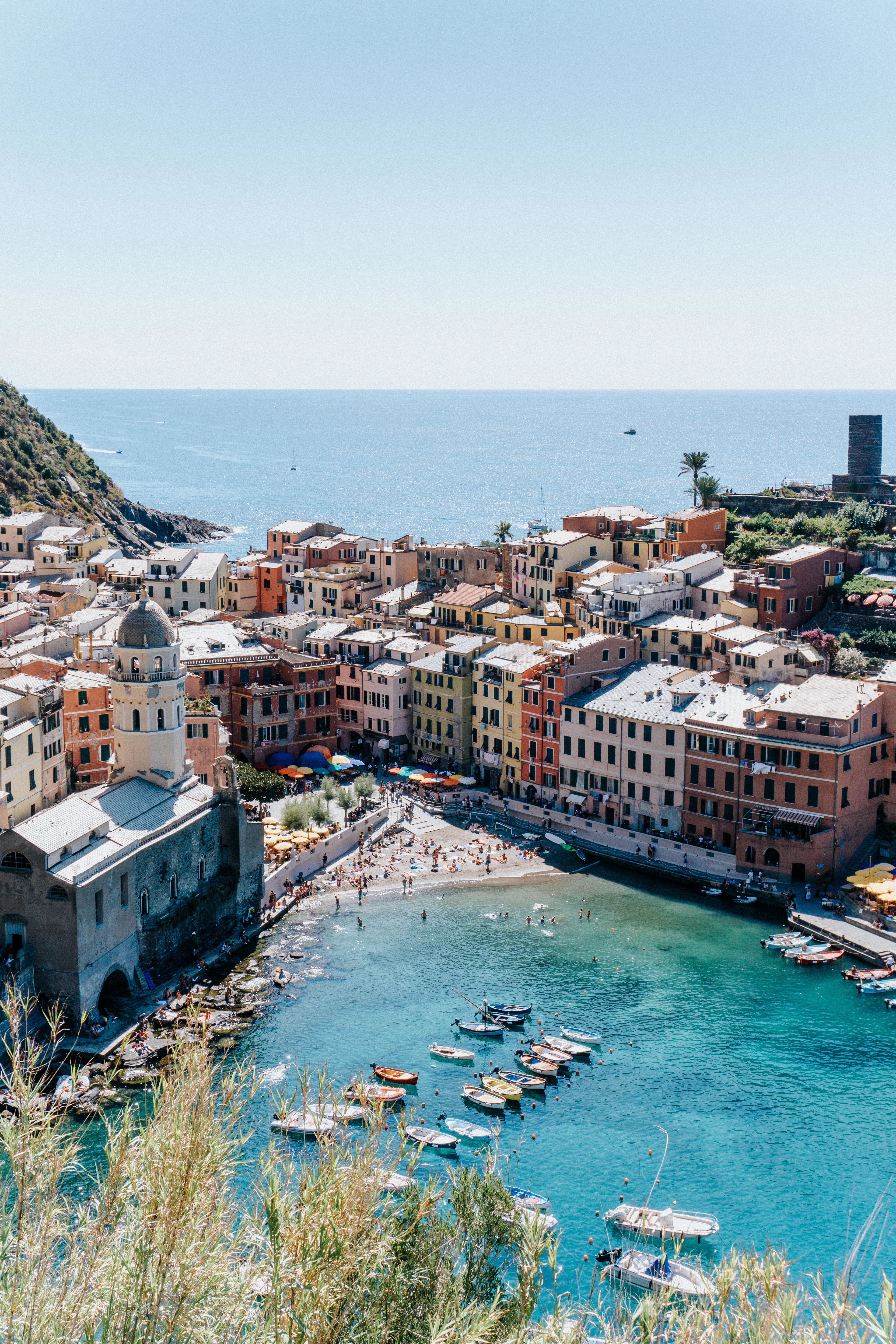 Vernazza's horseshoe harbour seen from the cliff path above, with pastel houses, a small beach and dinghies moored in clear turquoise water.