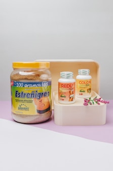 A collection of health supplement containers placed on a wooden tray with a decorative sprig of flowers. The large jar on the left displays a product labeled 'Estre&ntilde;igran', while two smaller bottles on the right are labeled 'REDU Gran Plus' and 'COLON Cleanser'. The setting has a light gray background with a lavender surface.