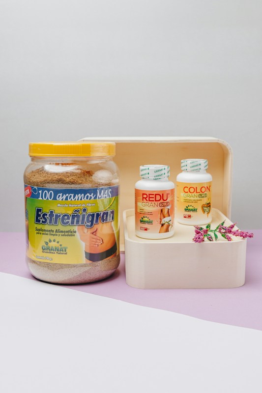 A collection of health supplement containers placed on a wooden tray with a decorative sprig of flowers. The large jar on the left displays a product labeled 'Estre&ntilde;igran', while two smaller bottles on the right are labeled 'REDU Gran Plus' and 'COLON Cleanser'. The setting has a light gray background with a lavender surface.
