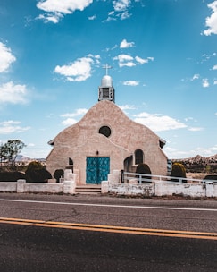 A small, rustic church with beige stucco walls and a prominent blue front door. A cross sits on top of the church's rounded roof, and a clear blue sky with scattered clouds serves as the backdrop. The foreground features a two-lane road, and the setting appears to be rural, with sparse vegetation and distant hills.