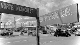 Street signs indicating Nortey Nyanchi Street and Nii Bonne Crescent are prominently visible. There is a road intersection with vehicles, including cars and a motorcycle, traversing. Telephone poles and wires are evident, and buildings, possibly residential or small businesses, line the street.