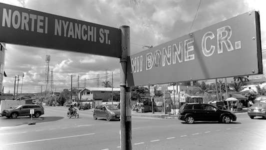 Street signs indicating Nortey Nyanchi Street and Nii Bonne Crescent are prominently visible. There is a road intersection with vehicles, including cars and a motorcycle, traversing. Telephone poles and wires are evident, and buildings, possibly residential or small businesses, line the street.