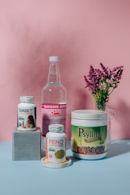 A collection of health supplement products is arranged against a pink backdrop. The items include bottles and containers labeled Diabet Gran Plus, Feno Gran Plus, Magne-Gran, and Gran Psyllium. A sprig of pink flowers in a glass vase adds a touch of nature to the composition.