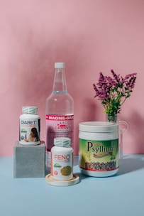 A collection of health supplement products is arranged against a pink backdrop. The items include bottles and containers labeled Diabet Gran Plus, Feno Gran Plus, Magne-Gran, and Gran Psyllium. A sprig of pink flowers in a glass vase adds a touch of nature to the composition.