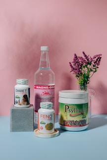 A collection of health supplement products is arranged against a pink backdrop. The items include bottles and containers labeled Diabet Gran Plus, Feno Gran Plus, Magne-Gran, and Gran Psyllium. A sprig of pink flowers in a glass vase adds a touch of nature to the composition.