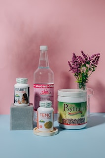 A collection of health supplement products is arranged against a pink backdrop. The items include bottles and containers labeled Diabet Gran Plus, Feno Gran Plus, Magne-Gran, and Gran Psyllium. A sprig of pink flowers in a glass vase adds a touch of nature to the composition.