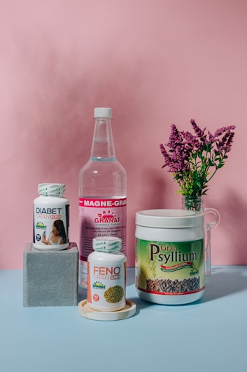 A collection of health supplement products is arranged against a pink backdrop. The items include bottles and containers labeled Diabet Gran Plus, Feno Gran Plus, Magne-Gran, and Gran Psyllium. A sprig of pink flowers in a glass vase adds a touch of nature to the composition.