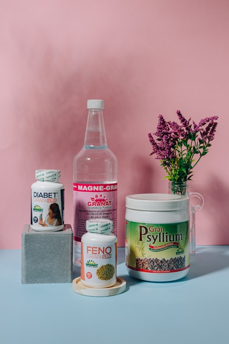 A collection of health supplement products is arranged against a pink backdrop. The items include bottles and containers labeled Diabet Gran Plus, Feno Gran Plus, Magne-Gran, and Gran Psyllium. A sprig of pink flowers in a glass vase adds a touch of nature to the composition.