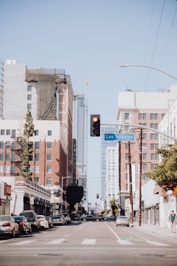 A city street is lined with tall buildings, some under construction with cranes visible in the distance. Several cars are parked along the sides, and traffic lights are visible, with a red light clearly illuminated. Trees and street signs are evident, including a sign indicating Los Angeles.