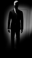 A faceless mannequin is dressed in a formal suit, standing against a backdrop of low lighting, creating a dramatic contrast between light and shadow.