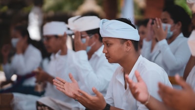 A group of people dressed in white are engaged in a ceremonial or prayer gathering. Many have their hands raised in a gesture of prayer or meditation. Some attendees wear traditional headpieces, and a few are wearing face masks.