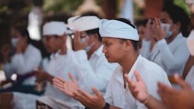 A group of people dressed in white are engaged in a ceremonial or prayer gathering. Many have their hands raised in a gesture of prayer or meditation. Some attendees wear traditional headpieces, and a few are wearing face masks.